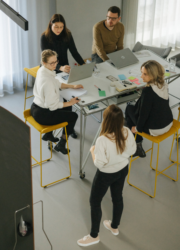Partnerin Petra Stahl mit drei Mitarbeiterinnen und einem Mitarbeiter im Creative Space um einen Tisch herumsitzend und -stehend. Das Bild ist von oben aus der Vogelperspektive aufgenommen.