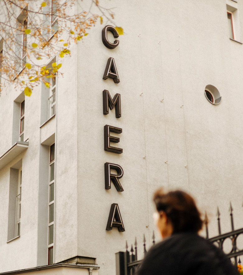 Das Bild zeigt das Gebäude von außen, in dem sich das Büro von Script befindet, das ehemalige Kino „Camera“ mit gleichem Schriftzug an der Seite. Im unscharfen Vordergrund läuft eine Passantin am Gebäude vorbei.