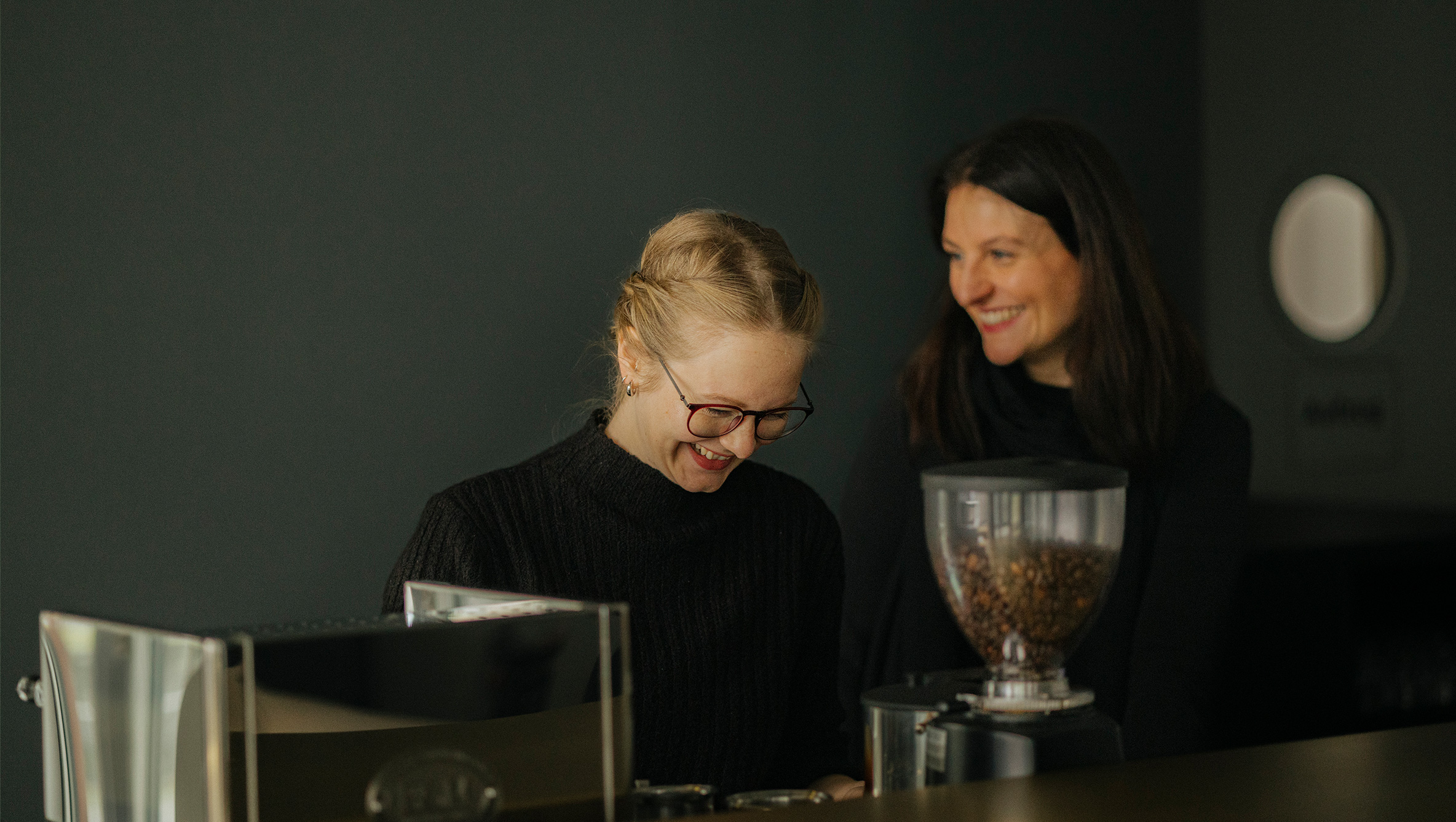 Das Foto zeigt zwei lachende Mitarbeiterinnen von Script hinter der Bar im Eingangsbereich der CAMERA. Eine von ihnen bereitet gerade Kaffee zu, die andere steht daneben.