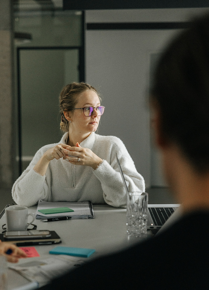 Aufnahme über die Schulter eines Mitarbeiters mit Fokus auf die gegenüber sitzende Mitarbeiterin. Sie richtet ihren Blick auf eine Person außerhalb des Bildes. Sie hat ihre Ellbogen auf dem Tisch, die Hände berühren sich und vor ihr liegen Schreibutensilien. Auf dem Tisch stehen außerdem ein Laptop, ein Glas und eine Tasse.