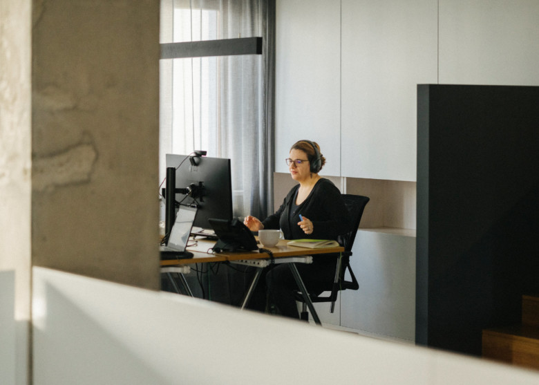 Eine Mitarbeiterin sitzt am Schreibtisch und schaut auf ihren Bildschirm. Sie trägt ein Headset. Das Foto ist aus der Distanz aufgenommen.